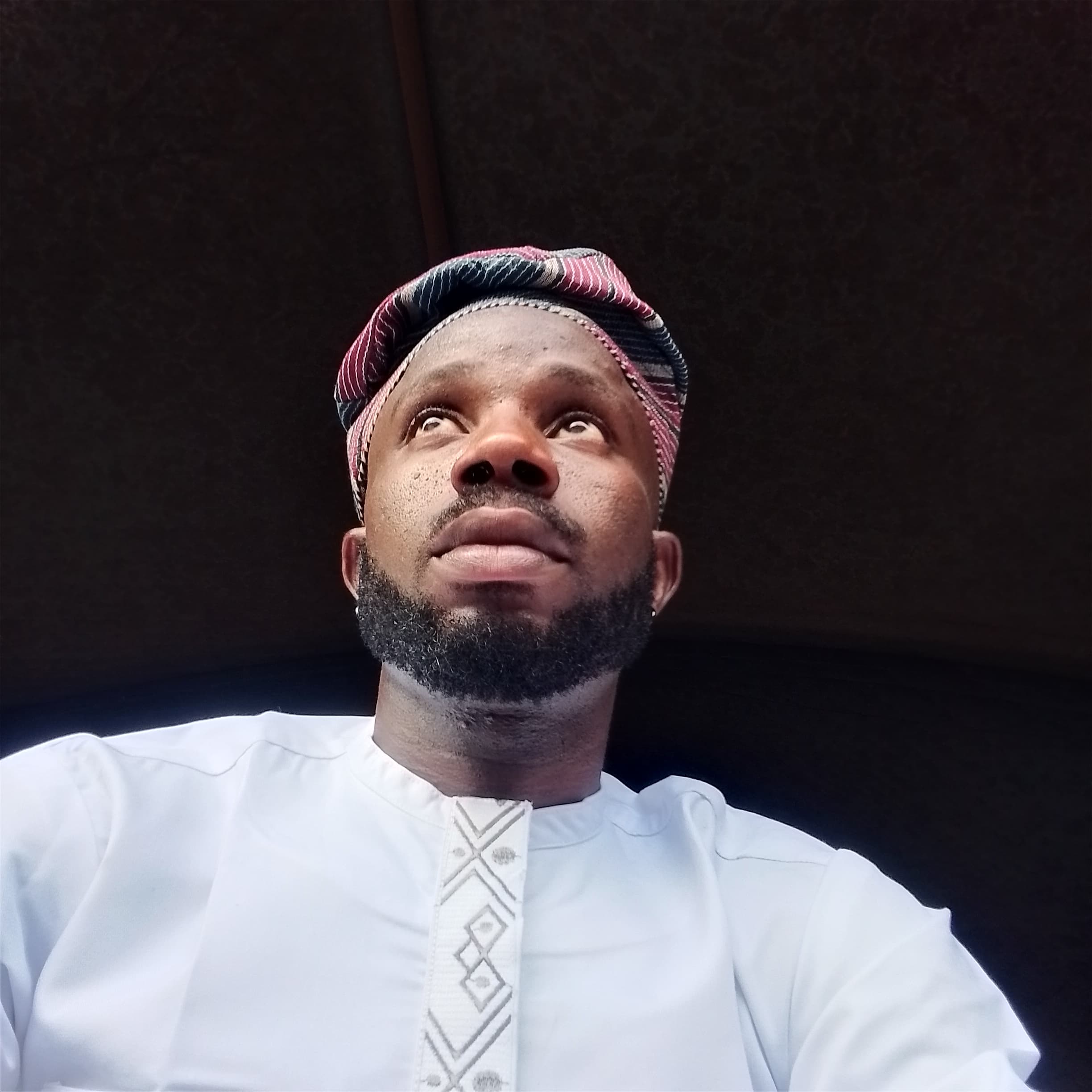 Bearded man in a patterned cap and white embroidered tunic gazing thoughtfully upwards.